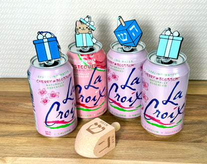 Four La Croix cans with Tab Buddies in different shades of blue resting atop the tabs on a wooden surface with a wooden dreidel displaying "shin" and "hay" on it.
