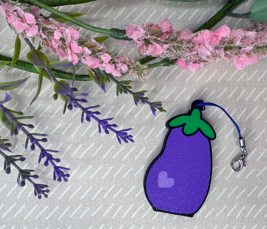Purple eggplant-shaped Tab Buddy keychain with green leaves on a beige background with pink and purple flowers.