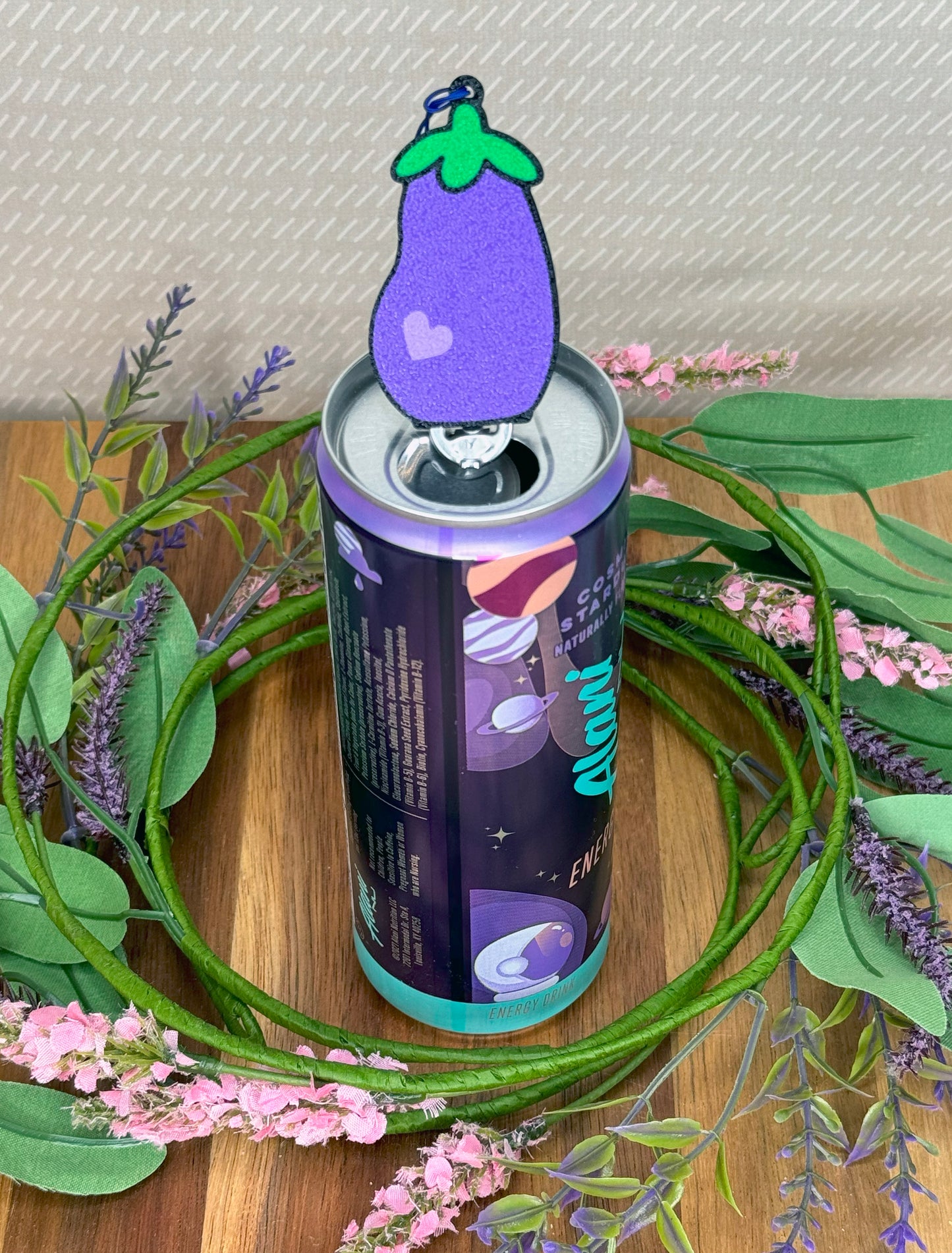 An energy drink can with a purple and green eggplant-shaped Tab Buddy on a wooden surface with greenery and flowers.