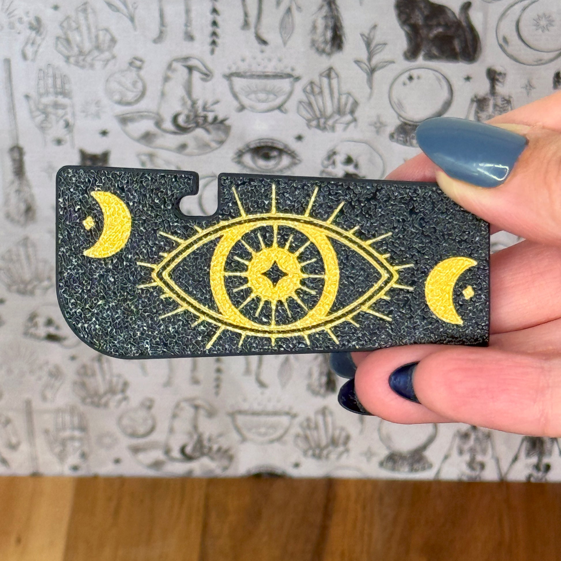 Hand holding a black and gold eye design Tab Buddy XL against a patterned background