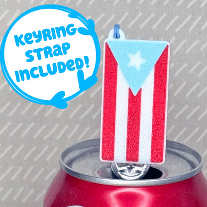A red, white and light blue Pureto Rico flag Tab Buddy soda can opener rests atop the tab of an open can of sparkling water with a beige background. Text reads, "Keyring strap included!"
