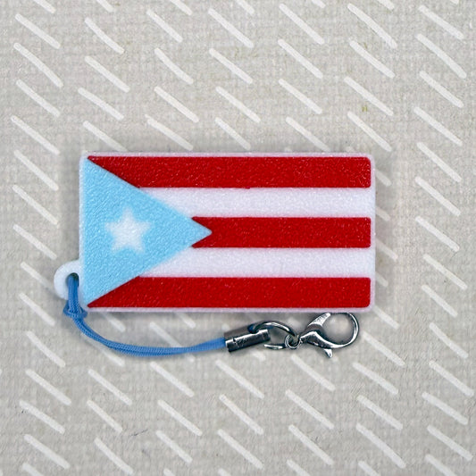 A red, white and light blue Pureto Rico flag Tab Buddy soda can opener rests atop a beige backgroun.
