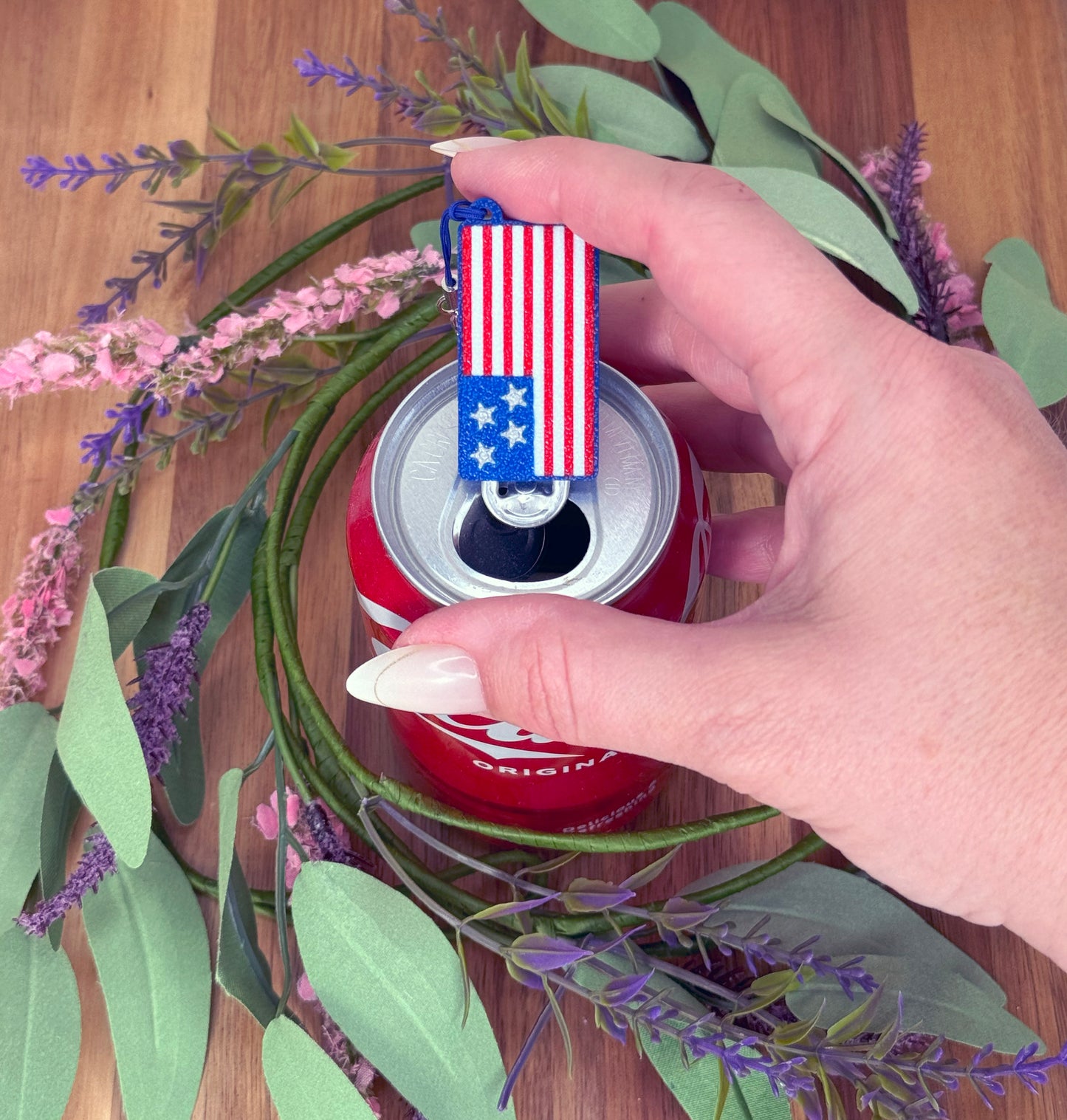 Hand holding a can of Coca-Cola with a USA flag Tab Buddy on top, surrounded by flowers and leaves.