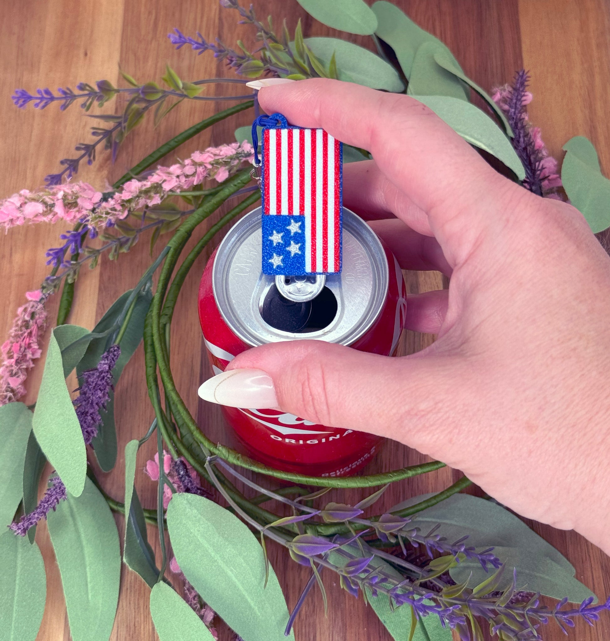 Hand holding a can of Coca-Cola with a USA flag Tab Buddy on top, surrounded by flowers and leaves.