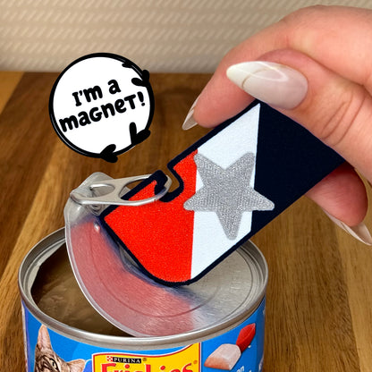 A hand holds a red, white and black flag-themed Tab Buddy XL as it opens a can of cat food on a wooden table. A speech bubble says 'I'm a magnet!'.