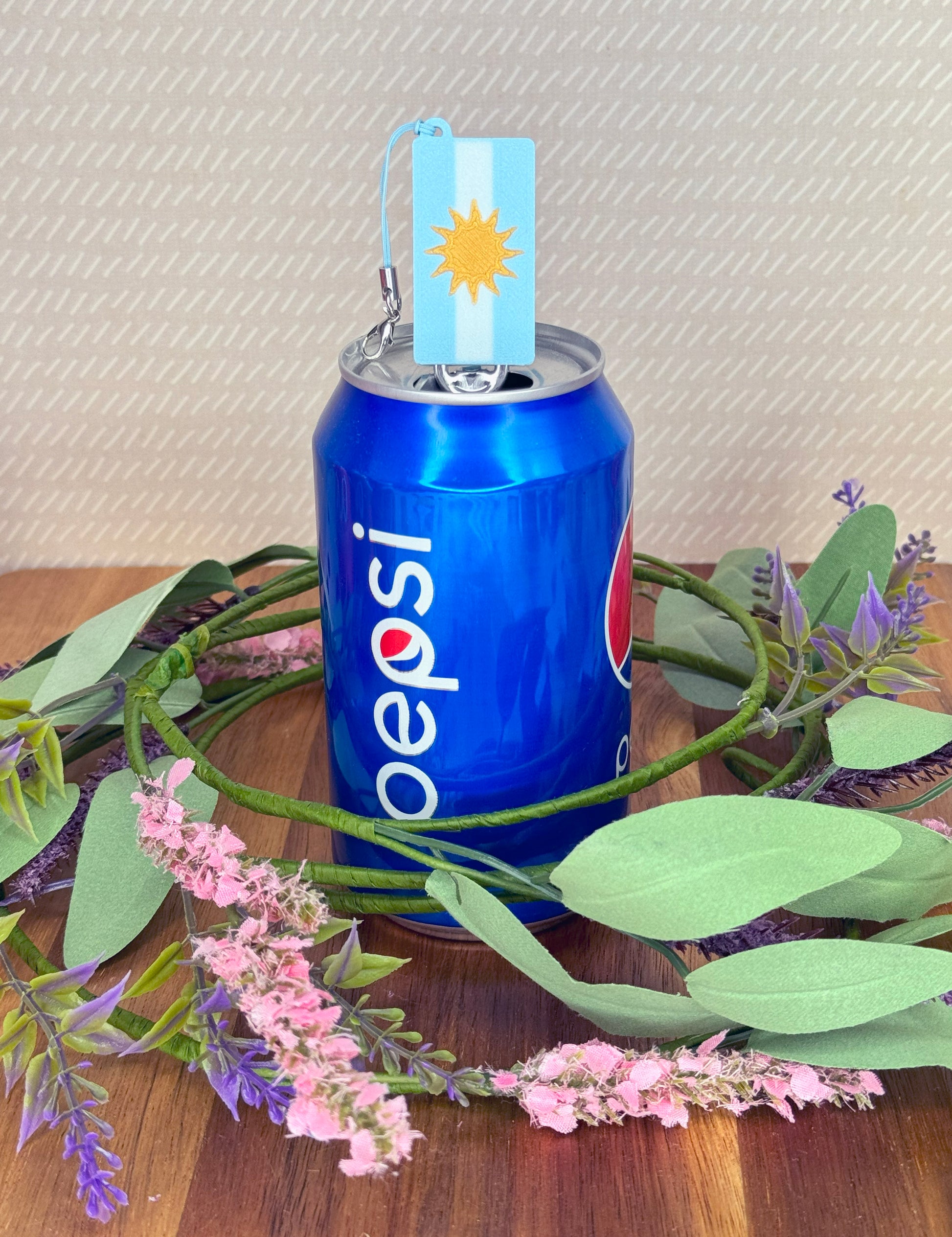 Pepsi can with a Argentina flag Tab Buddy, surrounded by greenery and flowers on a wooden surface.