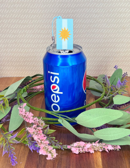 Pepsi can with a Argentina flag Tab Buddy, surrounded by greenery and flowers on a wooden surface.