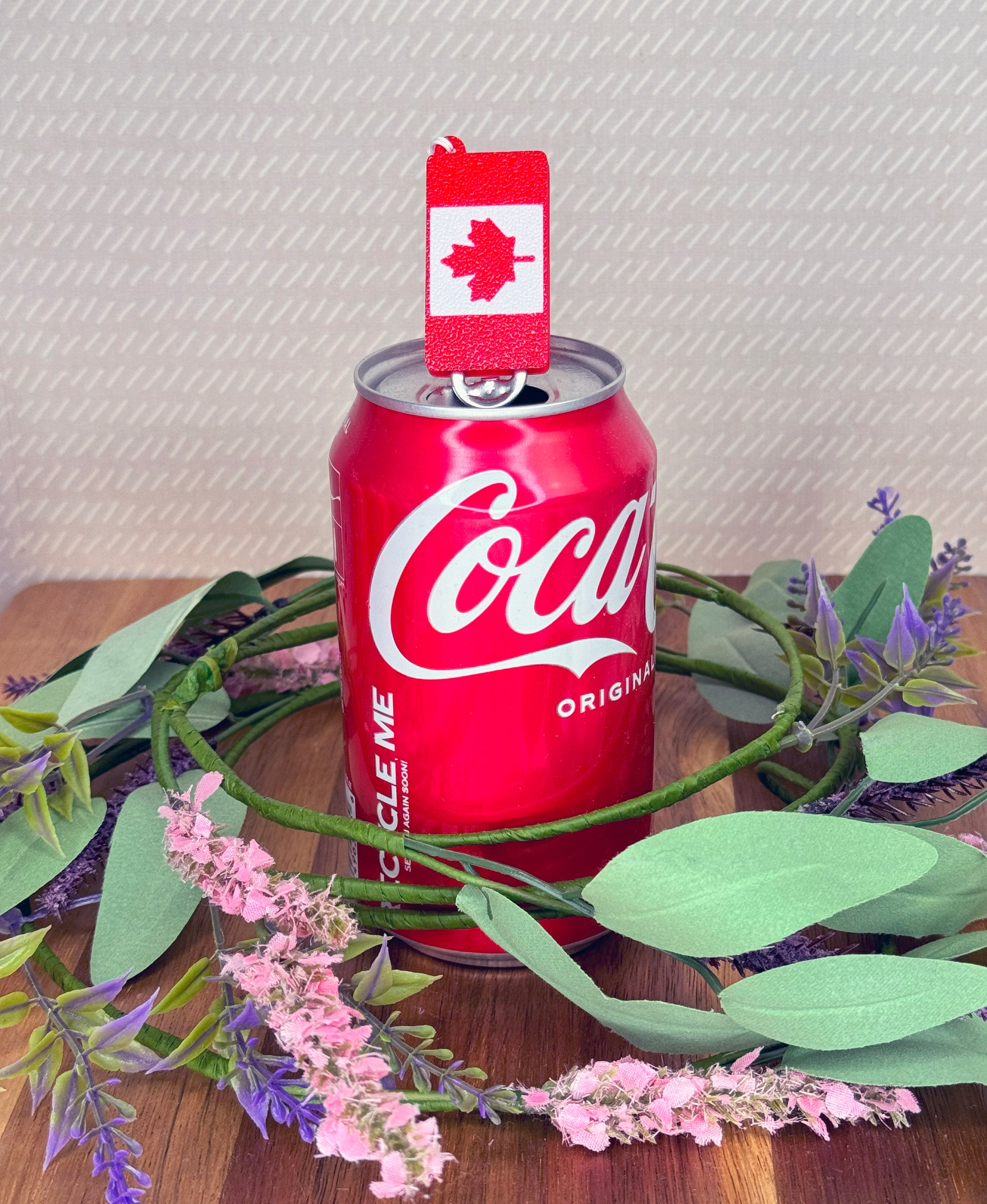 Red Coca-Cola can with a small Canada flag Tab Buddy on top, surrounded by greenery and flowers on a wooden surface.