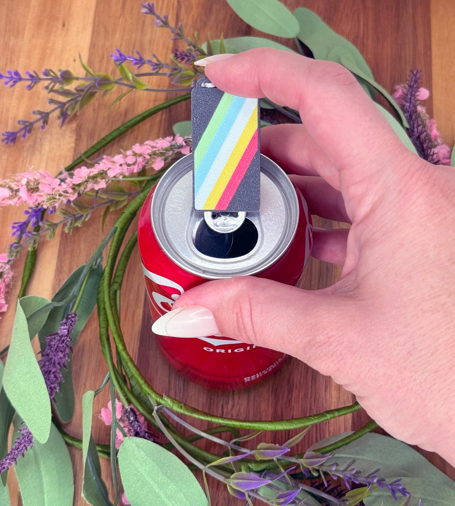 Disability pride flag Tab Buddy sits atop a can, surrounded by flowers on a wooden surface.
