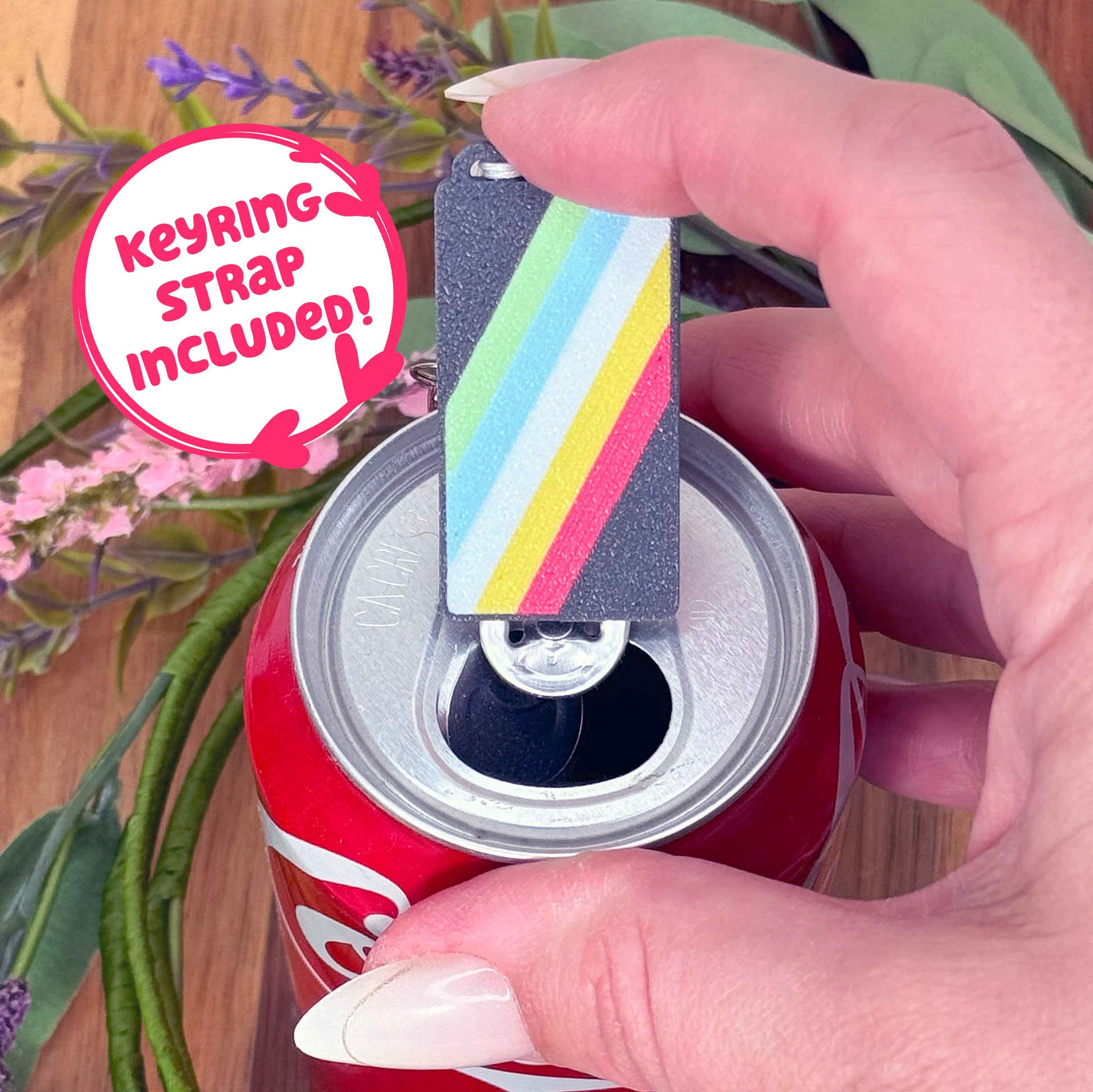 Hand holding a disability pride flag Tab Buddy with keyring strap attached, on a wooden surface with plants.