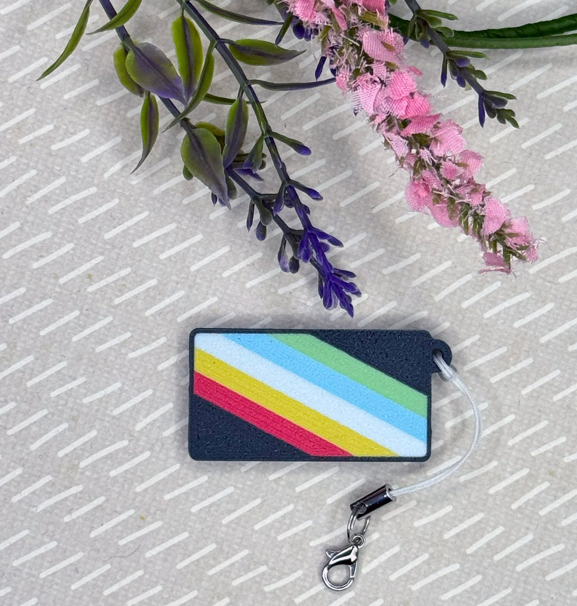 Tab Buddy with the disability pride flag on it with a strap attached rests on a beige surface with flowers.