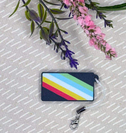 Tab Buddy with the disability pride flag on it with a strap attached rests on a beige surface with flowers.