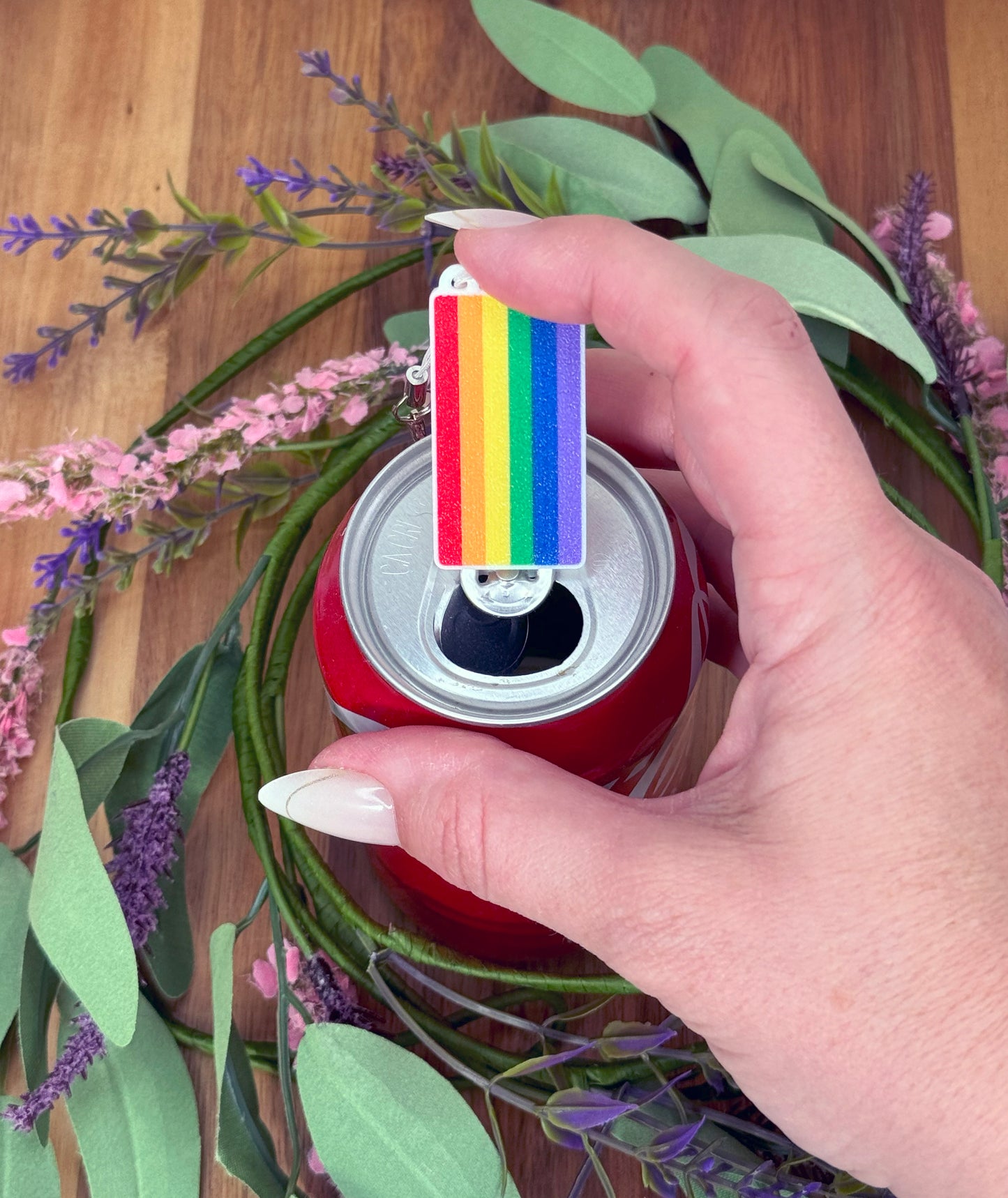 Hand holding a can with a rainbow-flag Tab Buddy against a floral background