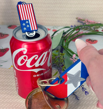 Red Coca-Cola can with a patriotic Tab Buddy keychain on a decorative surface. Tab Buddy XL in foreground opens a can of cat food.