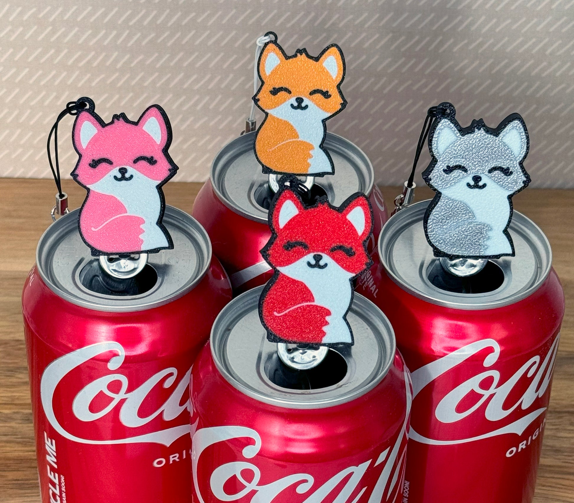 Four Coca-Cola cans with fox-shaped Tab Buddies in orange, pink, red and silver on a wooden surface.
