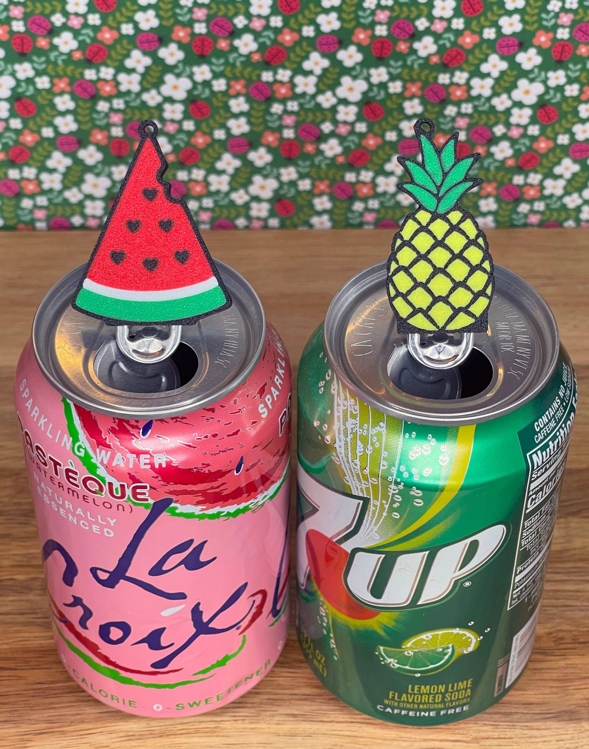 Two soda cans with watermelon and pineapple Tab Buddies resting atop on a wooden surface with a floral background.