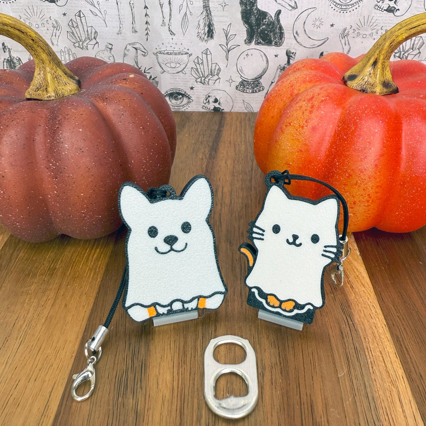 Two white ghost-themed Tab Buddies -- one a ghost cat and one a ghost dog -- on a wooden surface with pumpkins in the background.