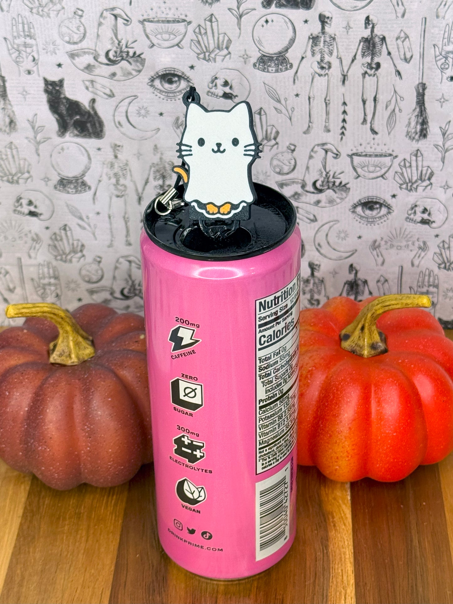 Pink can with a cat-shaped Tab Buddy keychain on top, surrounded by pumpkins against a patterned background