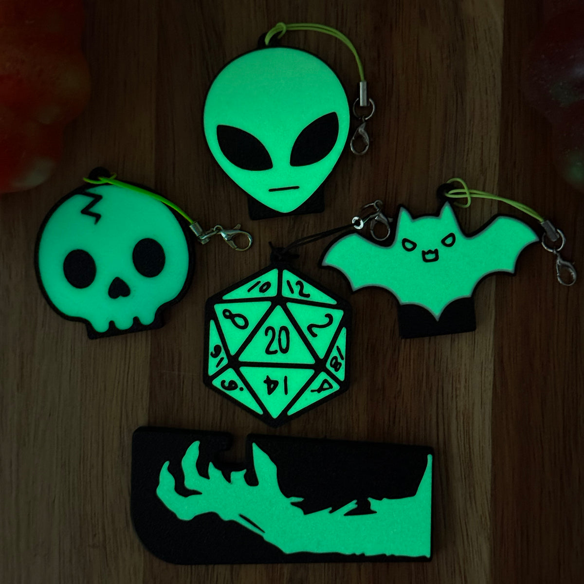 A set of five glow-in-the-dark Tab Buddies rest on a wooden surface at night. The designs include Soda Tab Buddies in the sahpe of an alien head, a skull, a 20-sided die, and a bat, and a Tab Buddy XL with a glowing zombie hand.