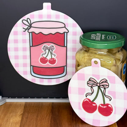 Two silicone Grippy Pads with cherry jam-themed gingham designs on a jar and a black surface.