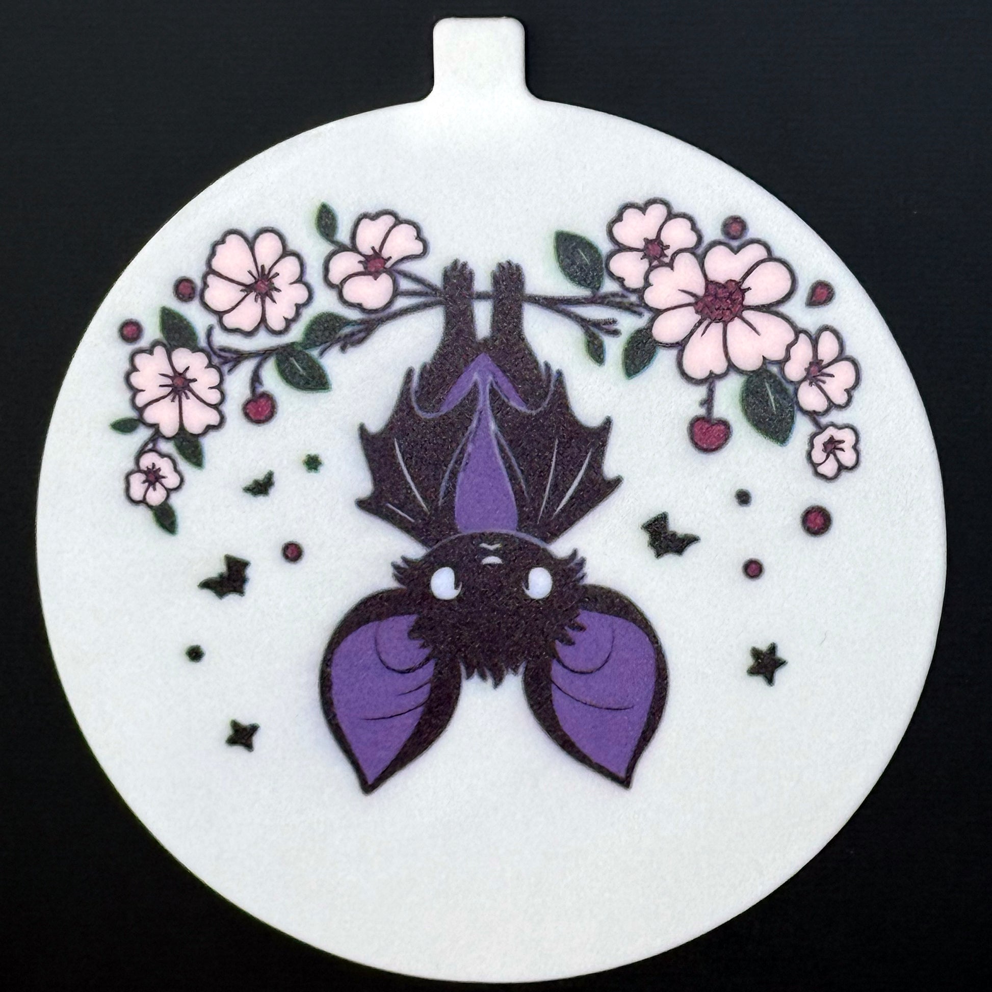 A circular Grippy Pad with a floral bat design hangs via its small internal magnet from a black surface.