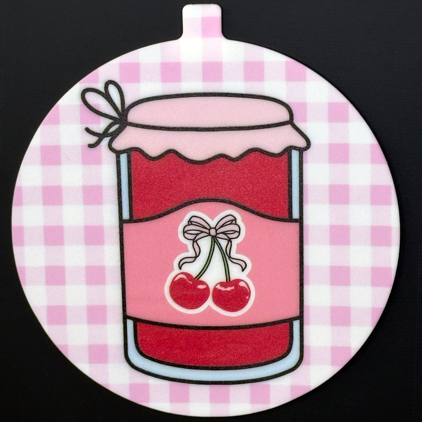 A circular Grippy Pad with a pink and red cherry jam design hangs via its small internal magnet from a black surface.