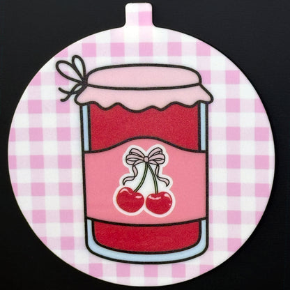 A circular Grippy Pad with a pink and red cherry jam design hangs via its small internal magnet from a black surface.