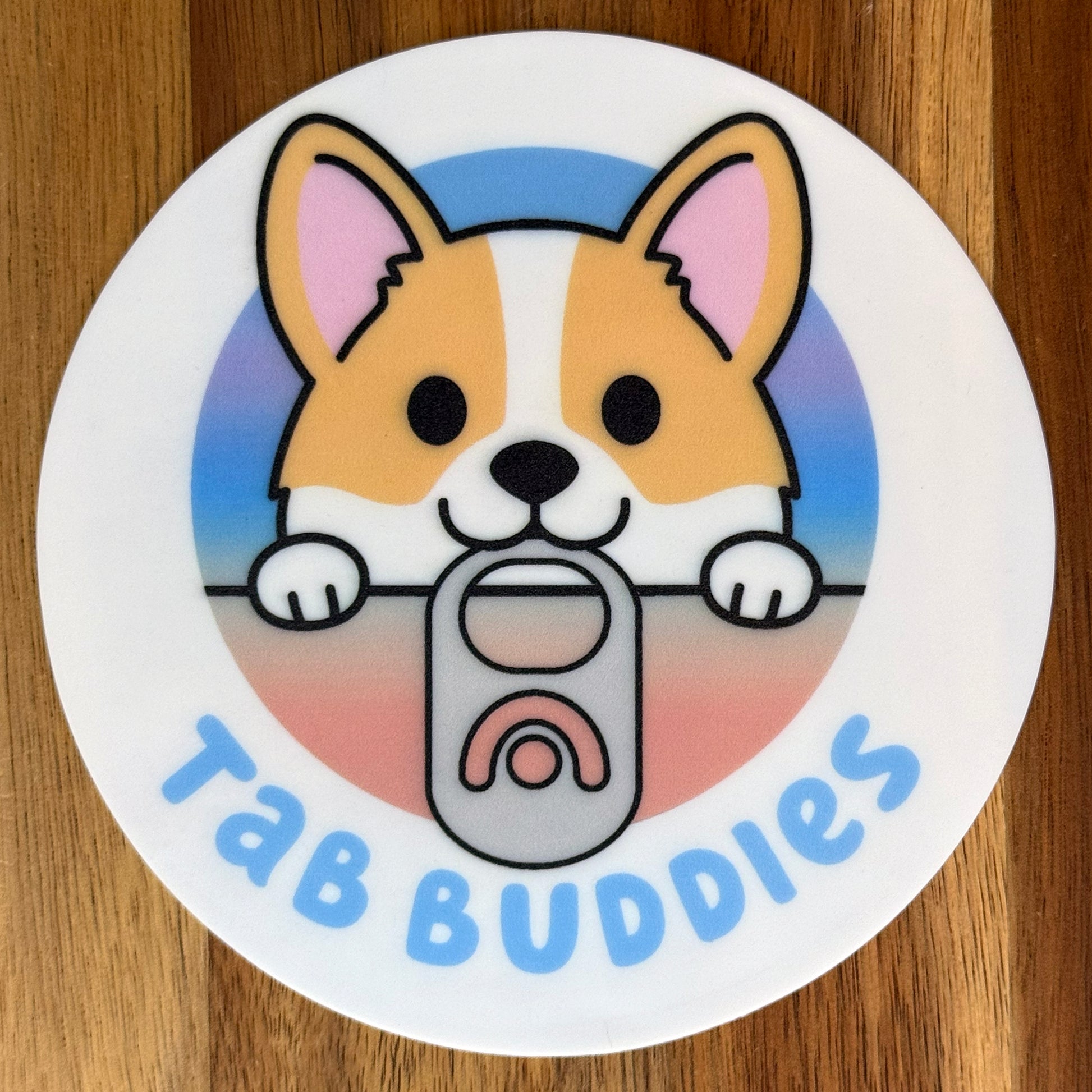 Circular grippy  pad with a cartoon corgi holding a tab and 'Tab Buddies' text on a wooden surface