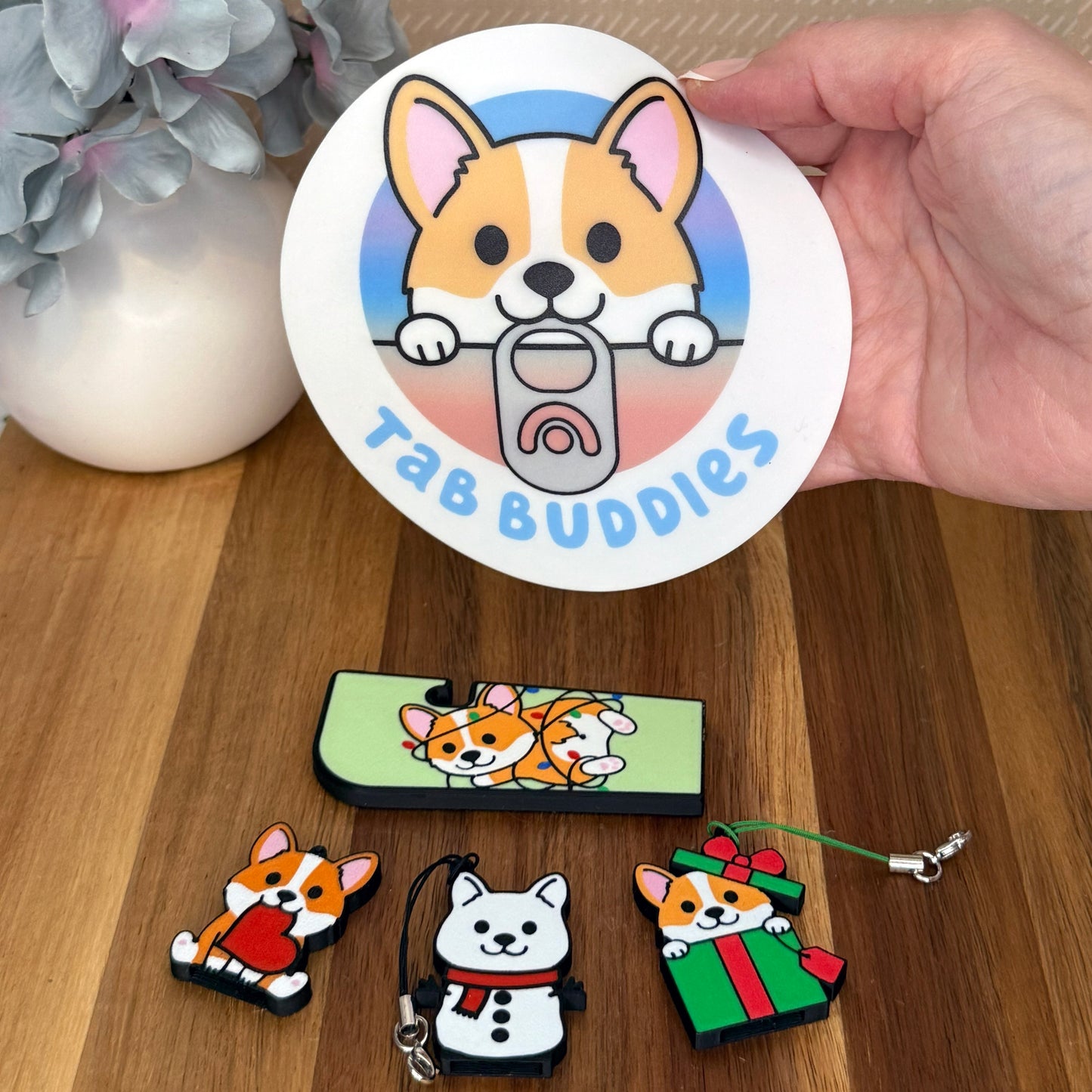 Collection of Tab Buddies corgi-themed product on a wooden surface with a hand holding a Corgi Grippy Pad.