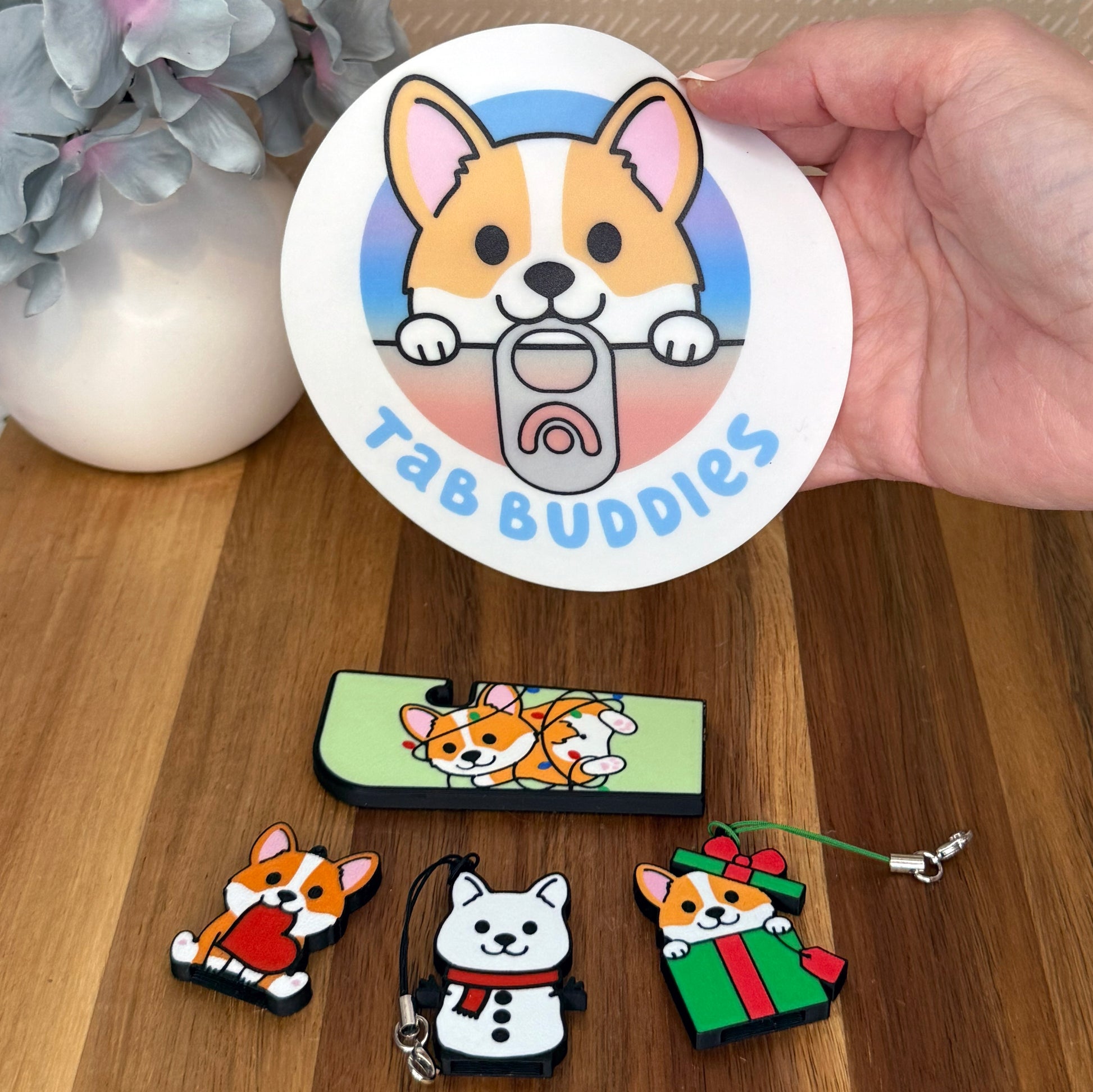 Collection of Tab Buddies corgi-themed product on a wooden surface with a hand holding a Corgi Grippy Pad.