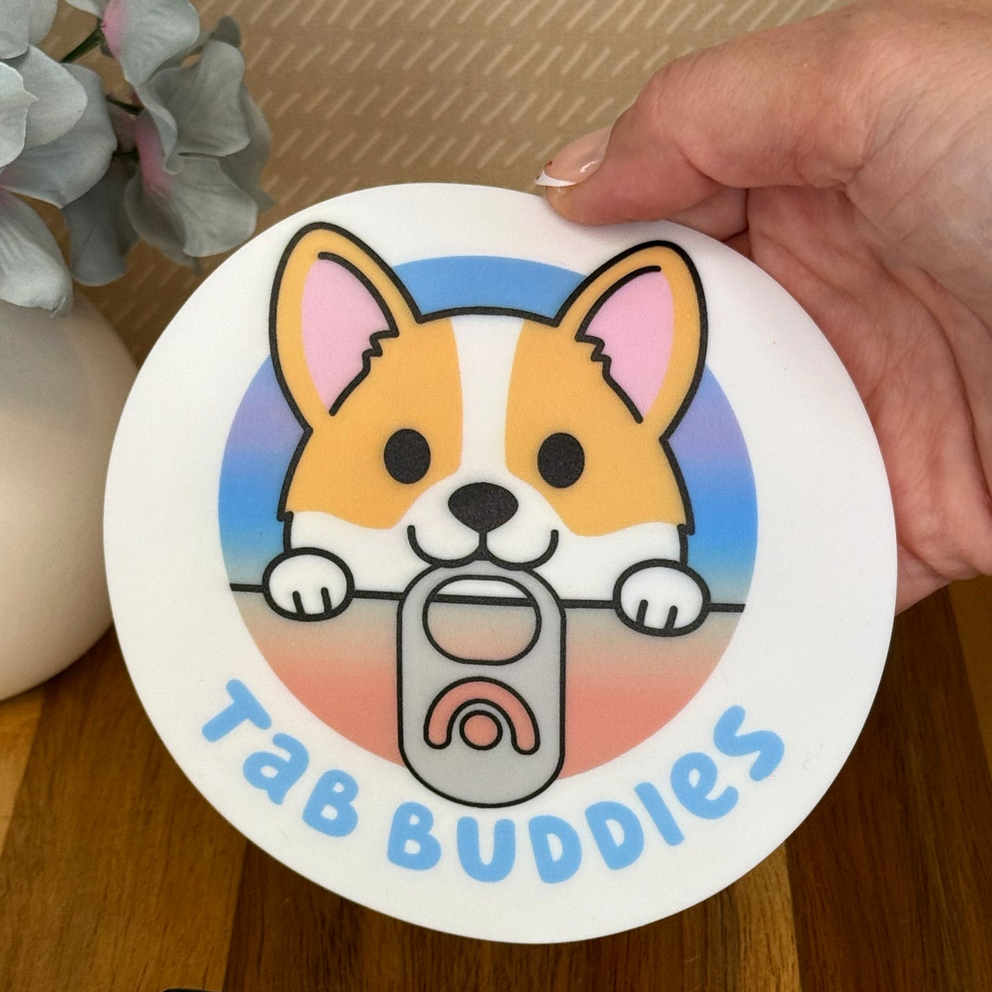 A hand holding a cartoon corgi design  grippy pad with 'Tab Buddies' text over a wooden surface
