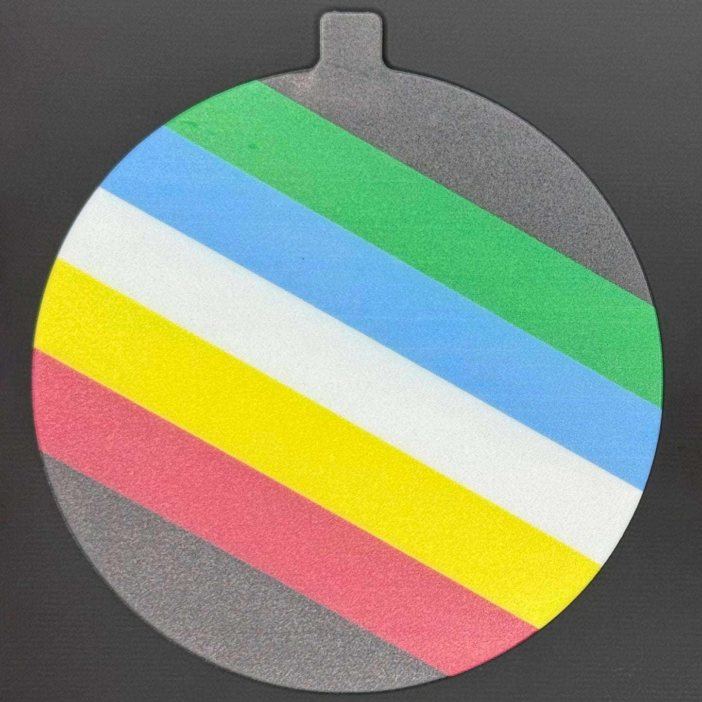 The large 5" diameter size Grippy Pad of the Disability Pride set is shown against a grey background. The design  includes an internal magnet in a tab at the top and features the Disability Pride flag spanning across the surface of the pad.