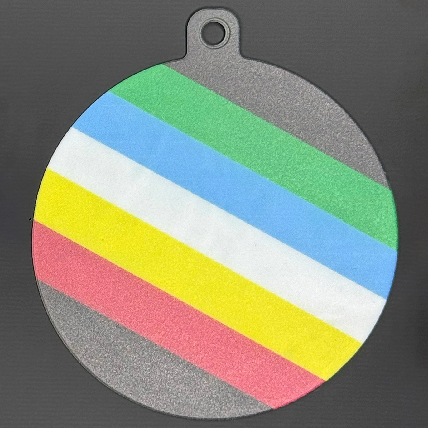 The small 3.5" diameter size Grippy Pad of the Disability Pride set is shown against a grey background. The design  includes a keychain hole set into a tab at the top and features the Disability Pride flag spanning across the surface of the pad.