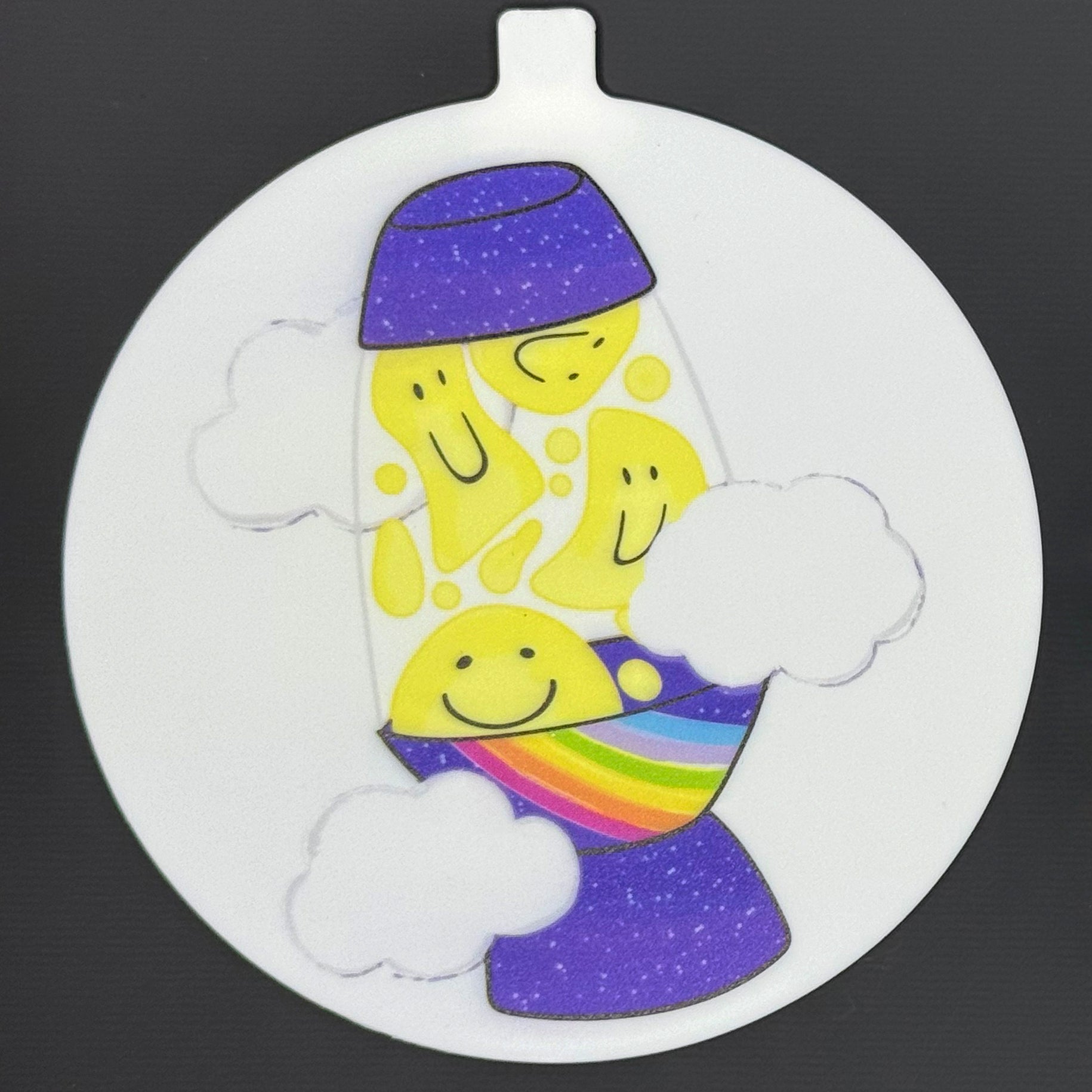 The large 5" diameter size Grippy Pad of the Groovy Pride set is shown against a grey background. The design  includes an internal magnet in a tab at the top and features a purple galaxy lava lamp with yellow smiley faces in the lava, a rainbow along one edge and clouds surrounding it.