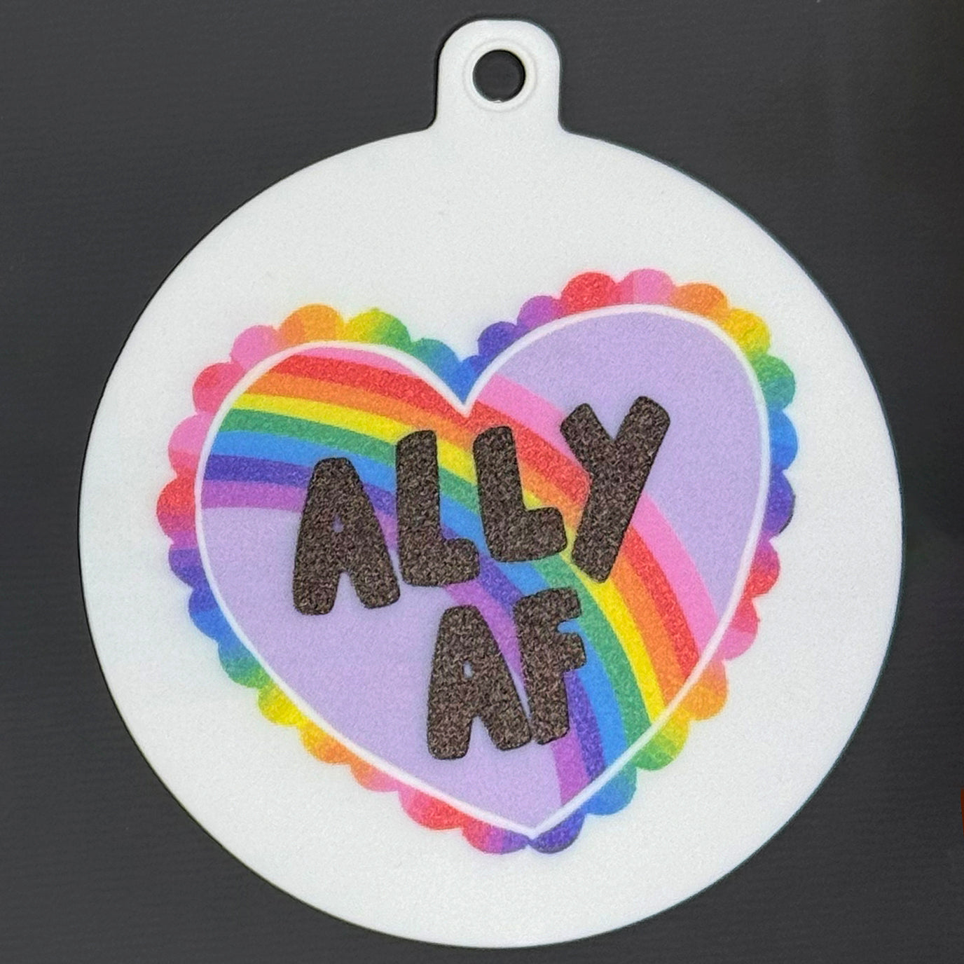 The small 3.5" diameter size Grippy Pad of the Groovy Pride set is shown against a grey background. The design  includes a keychain hole set into a tab at the top and features a colorful rainbow heart with scalloped edges and the text "Ally AF" in the center.