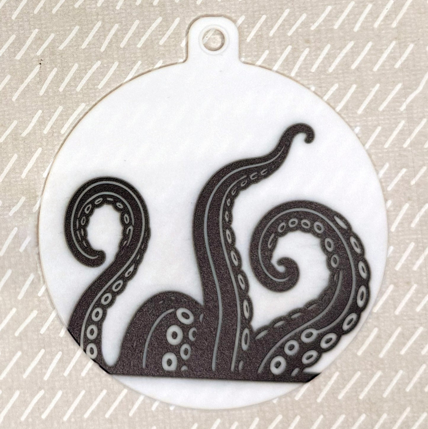 The small 3.5" diameter size Grippy Pad of theKraken Wave set is shown against a beige background. The design  includes a keychain hole set into a tab at the top and features kraken tentacles in black and white rising up from the bottom of the pad toward the top.