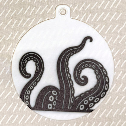 The small 3.5" diameter size Grippy Pad of theKraken Wave set is shown against a beige background. The design  includes a keychain hole set into a tab at the top and features kraken tentacles in black and white rising up from the bottom of the pad toward the top.