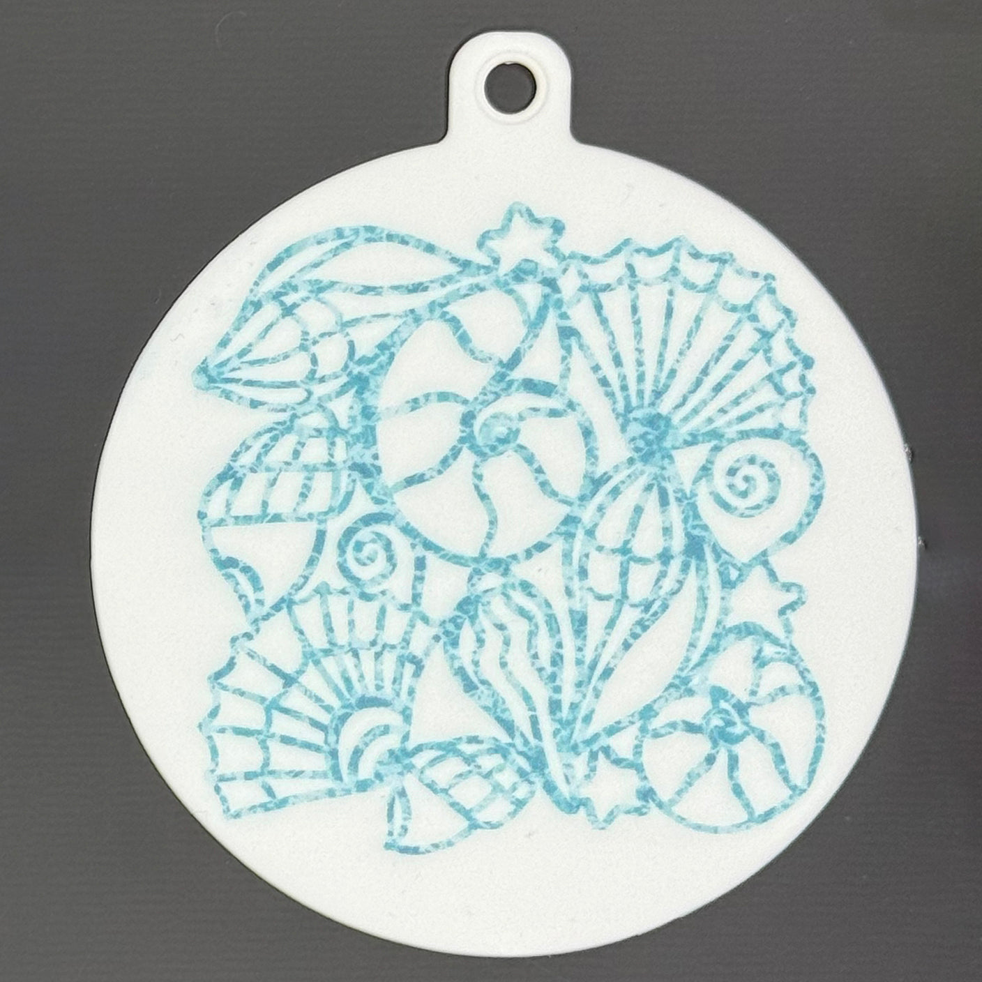 The small 3.5" diameter size Grippy Pad of the Sunny Shores set is shown against a grey background. The design  includes a keychain hole set into a tab at the top and features a design of aqua-colored shells sparkling on the surface.