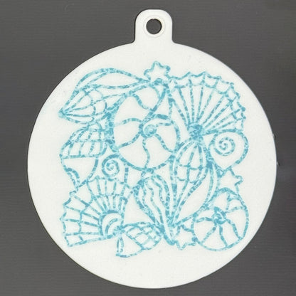 The small 3.5" diameter size Grippy Pad of the Sunny Shores set is shown against a grey background. The design  includes a keychain hole set into a tab at the top and features a design of aqua-colored shells sparkling on the surface.