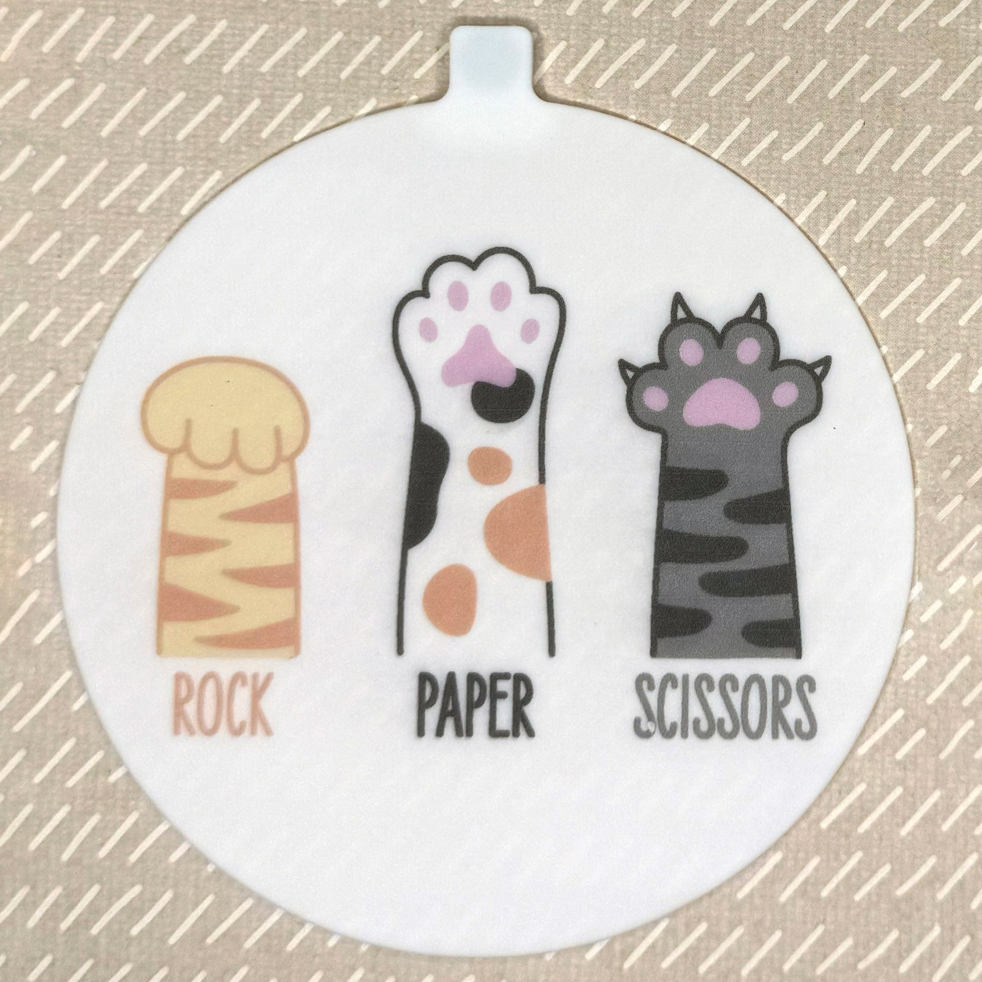 The large 5" diameter size Grippy Pad of the Rock Paper Scissors set is shown against a beige background. The design  includes an internal magnet in a tab at the top and features 3 cat paws with the words "Rock Paper Scissors" underneath in matching text.