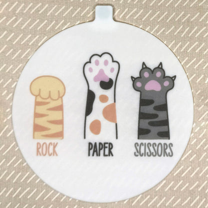 The large 5" diameter size Grippy Pad of the Rock Paper Scissors set is shown against a beige background. The design  includes an internal magnet in a tab at the top and features 3 cat paws with the words "Rock Paper Scissors" underneath in matching text.