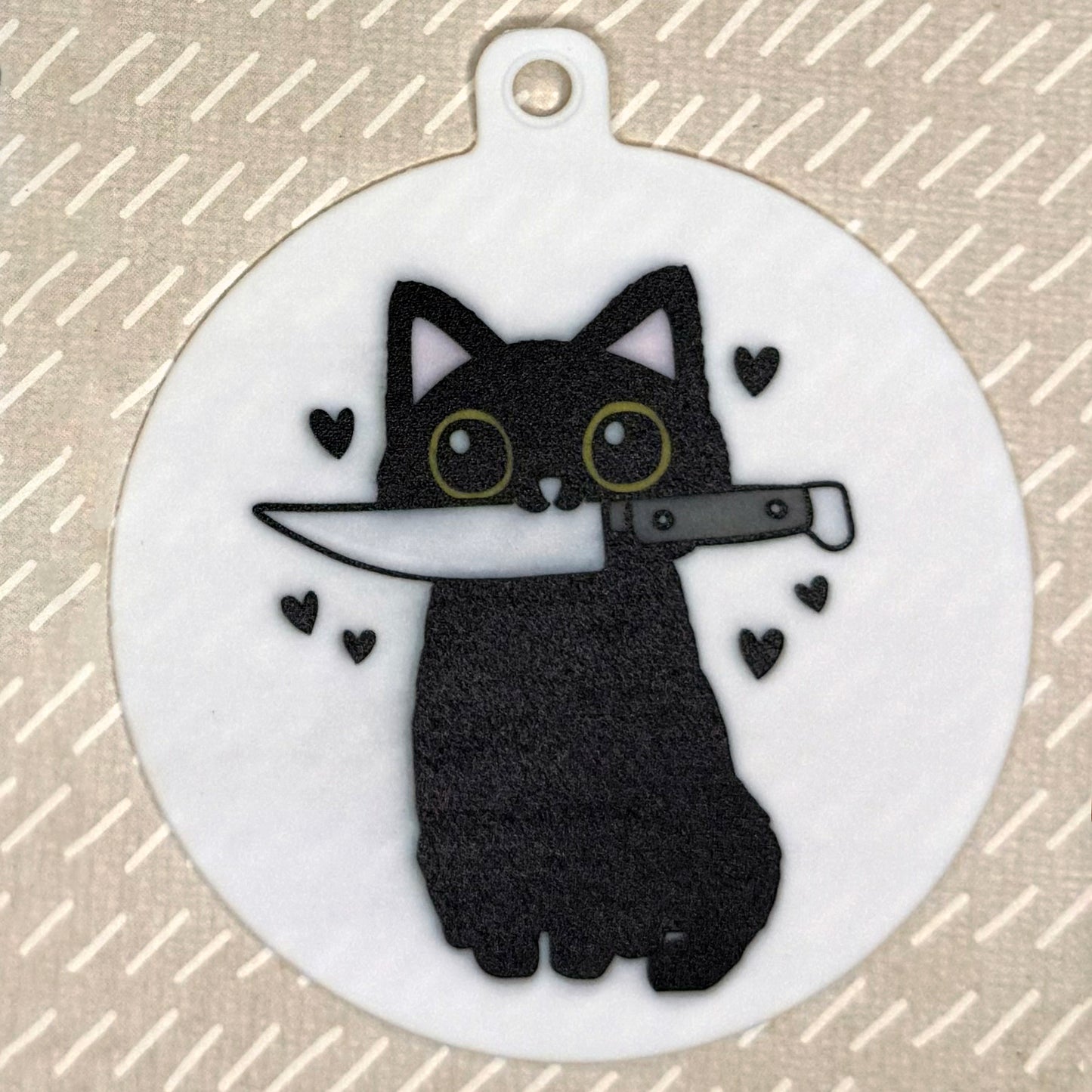 The small 3.5" diameter size Grippy Pad of the Paw Paper Scissors cat set is shown against a grey background. The design  includes a keychain hole set into a tab at the top and features a vlack cat holding a knife in its mouth with black hearts surrounding it.
