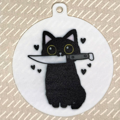 The small 3.5" diameter size Grippy Pad of the Paw Paper Scissors cat set is shown against a grey background. The design  includes a keychain hole set into a tab at the top and features a vlack cat holding a knife in its mouth with black hearts surrounding it.