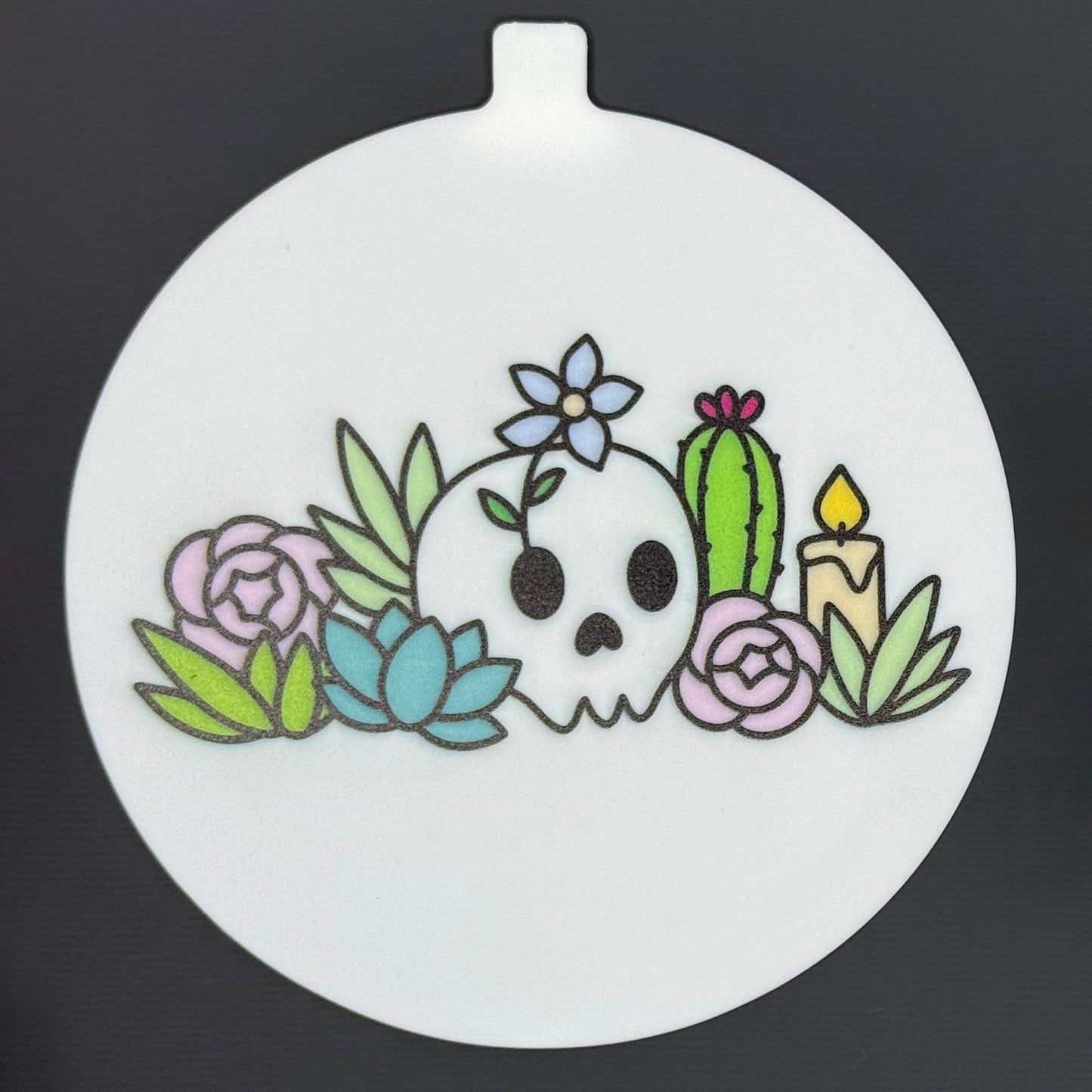 The large 5" diameter size Grippy Pad of the Mystic Garden set is shown against a grey background. The design  includes an internal magnet in a tab at the top and features a garden of cacti, succulents, a candle and flowers with a skull.