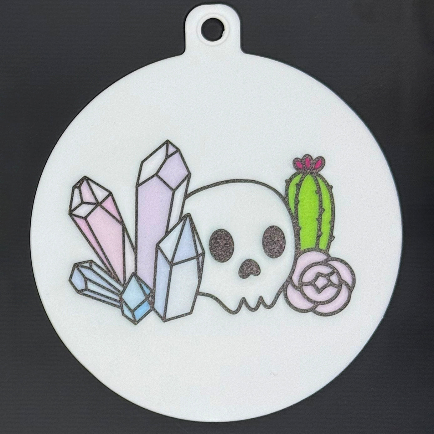 The small 3.5" diameter size Grippy Pad of the Mystic Garden set is shown against a grey background. The design  includes a keychain hole set into a tab at the top and features a skull, cactus, flower and colorful crystals.