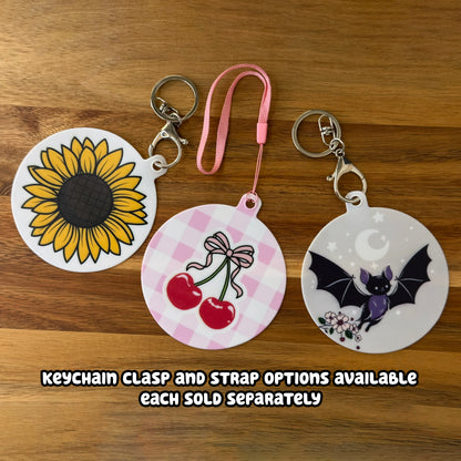 Three small circular Grippy Pads of various designs with keychains and strapped attached rest on a wooden surface. Text indicates, "Keychain clasp and strap options available. Each sold separately."