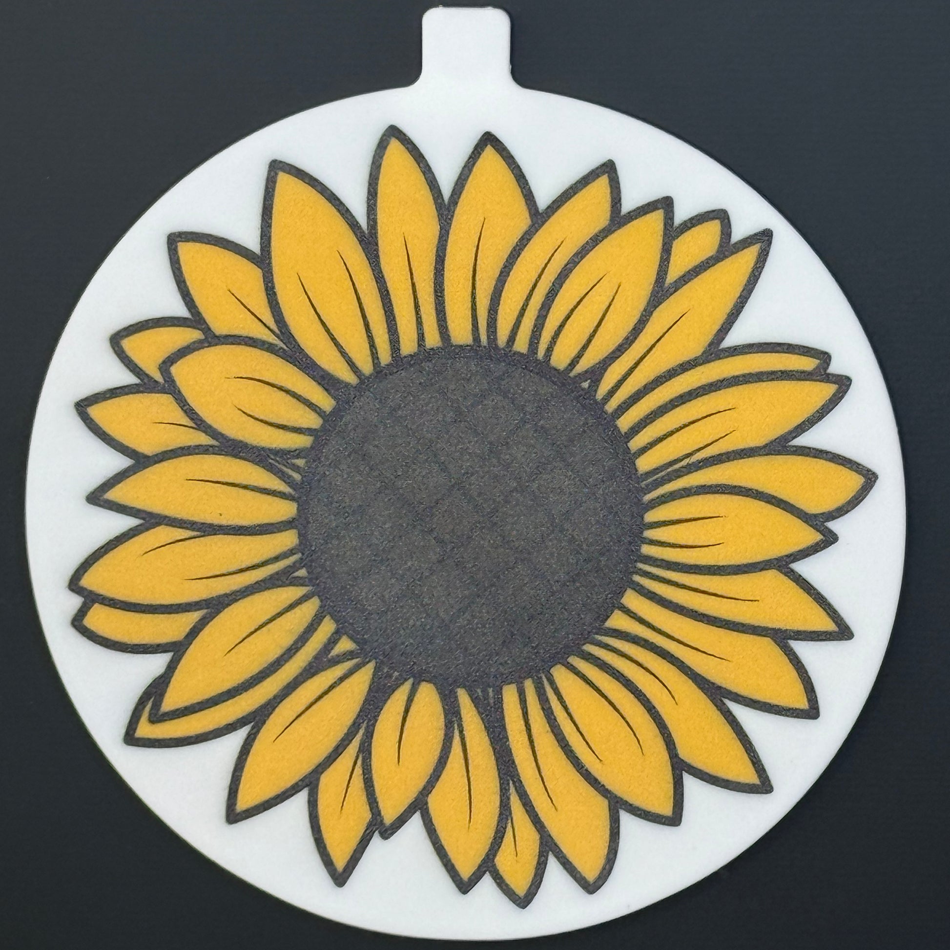 A circular Grippy Pad with a sunflower design hangs via its small internal magnet from a black surface.