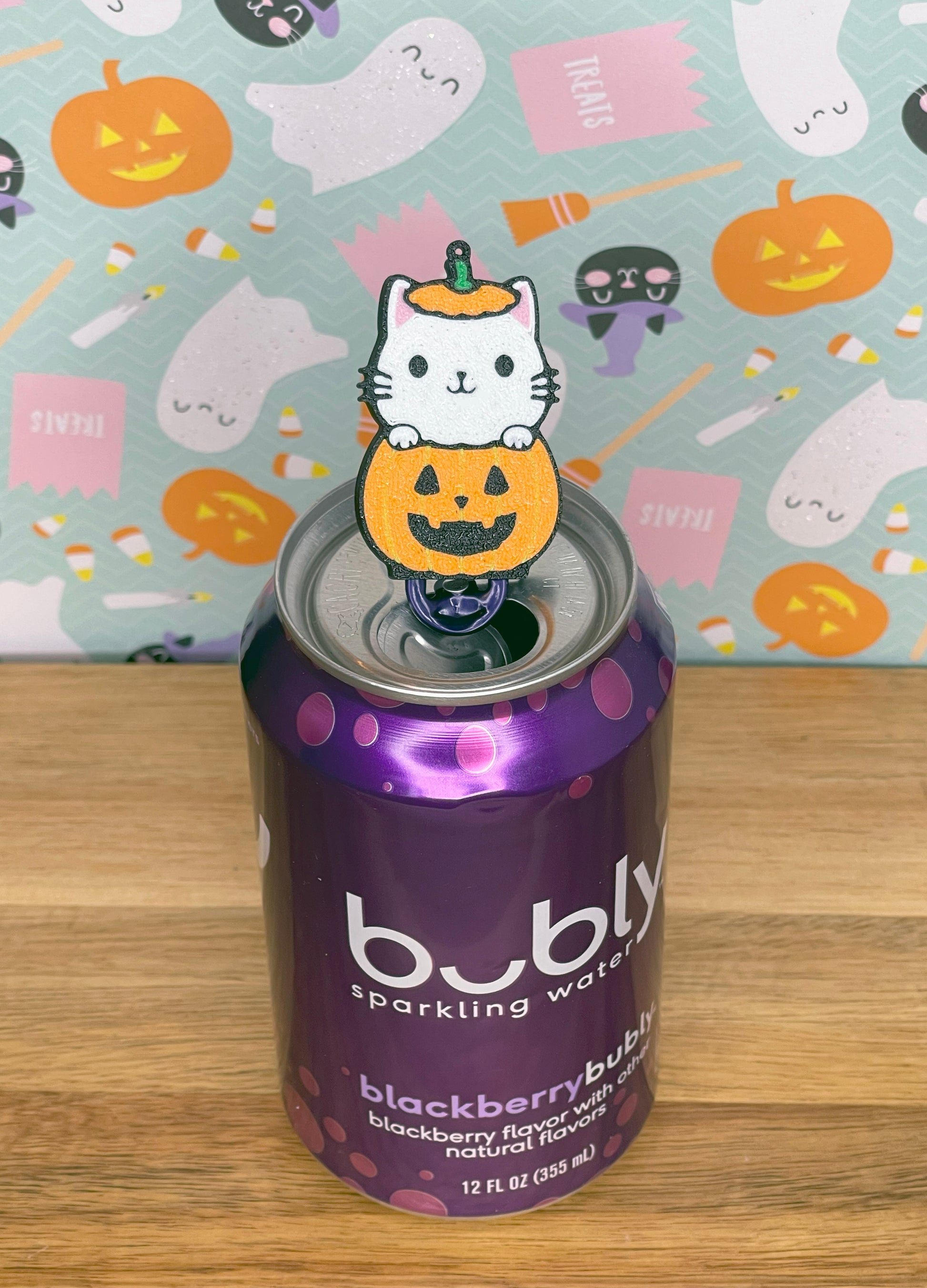 Bubly sparkling water can with a Halloween-themed white cat peeking out of a pumpkin Tab Buddy rests atop a colorful Halloween-themed background.