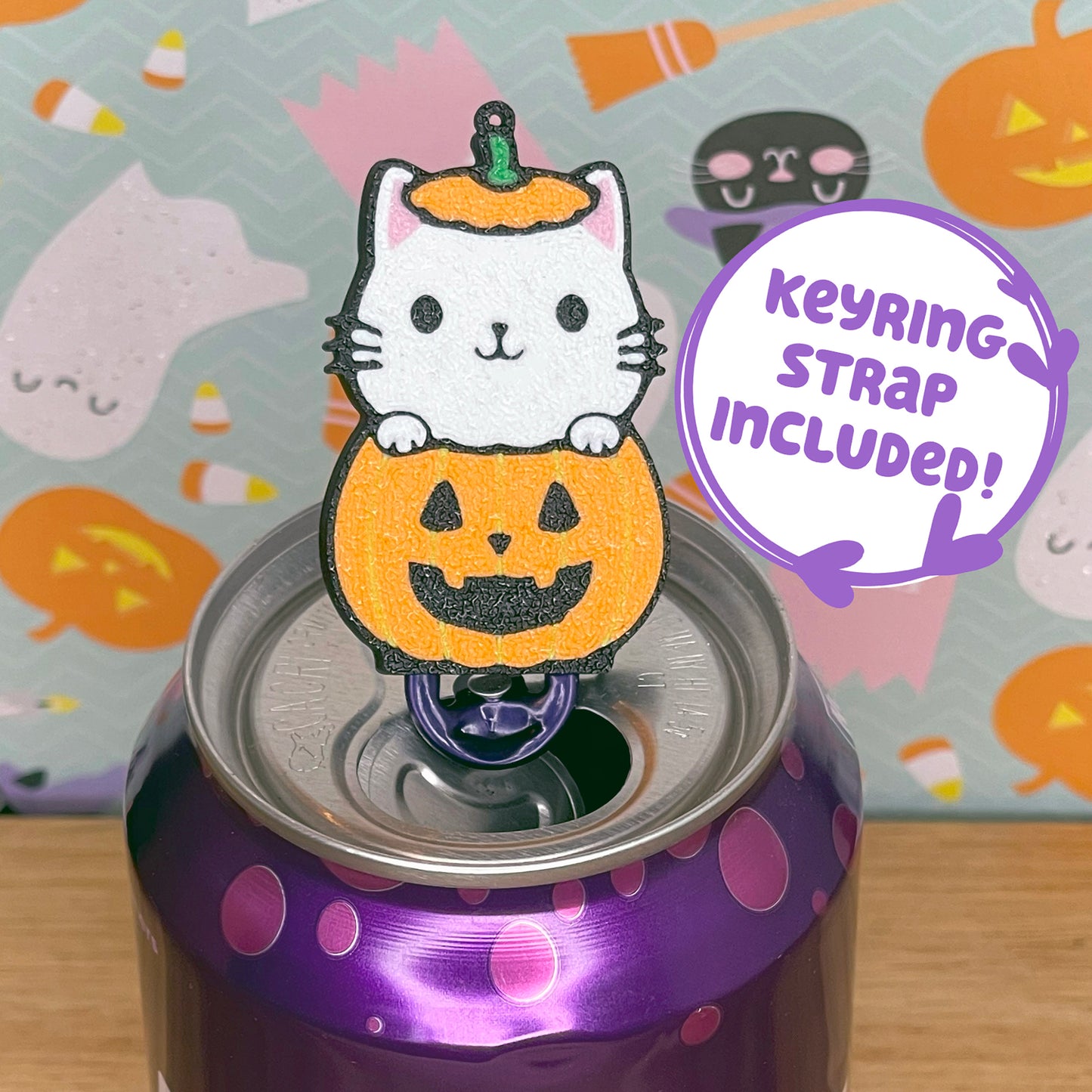 Halloween-themed cat-in-pumpkin Tab Buddy with keyring strap on a purple can with a colorful background featuring pumpkins and ghosts.