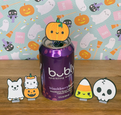 A sparkling water can with Halloween-themed Tab Buddies on a wooden surface and colorful Halloween-patterned background.
